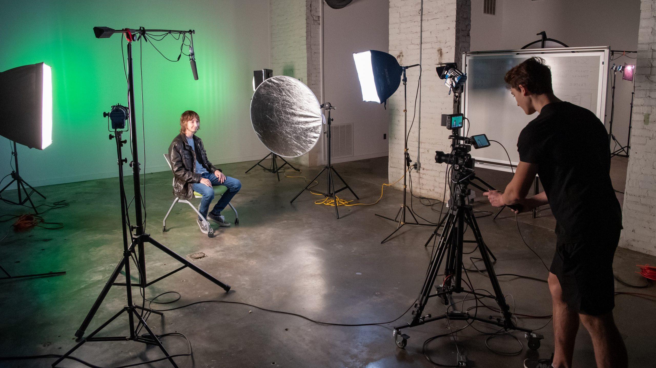 Student sitting in a chair with video equipment and lighting placed around him, while another student operates a camera on a tripod.